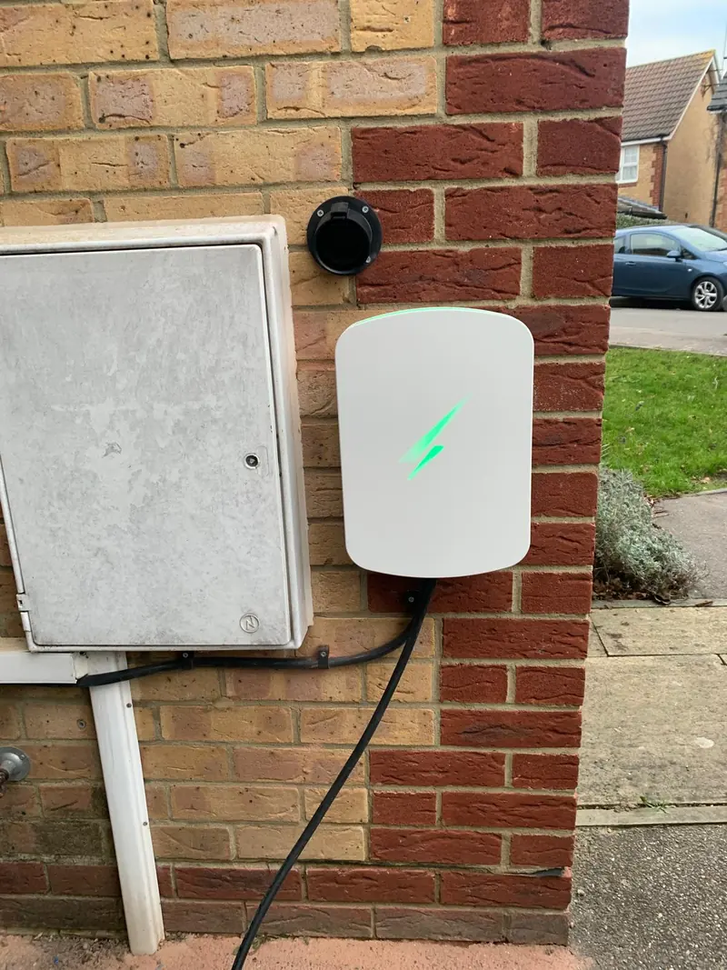 White wall-mounted EV charger with a green light, fixed to red brick beside a grey meter box