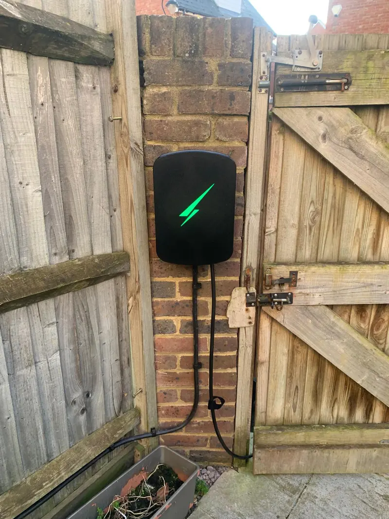 Black EV charger with a green light on a brick pier between a timber fence and a gate